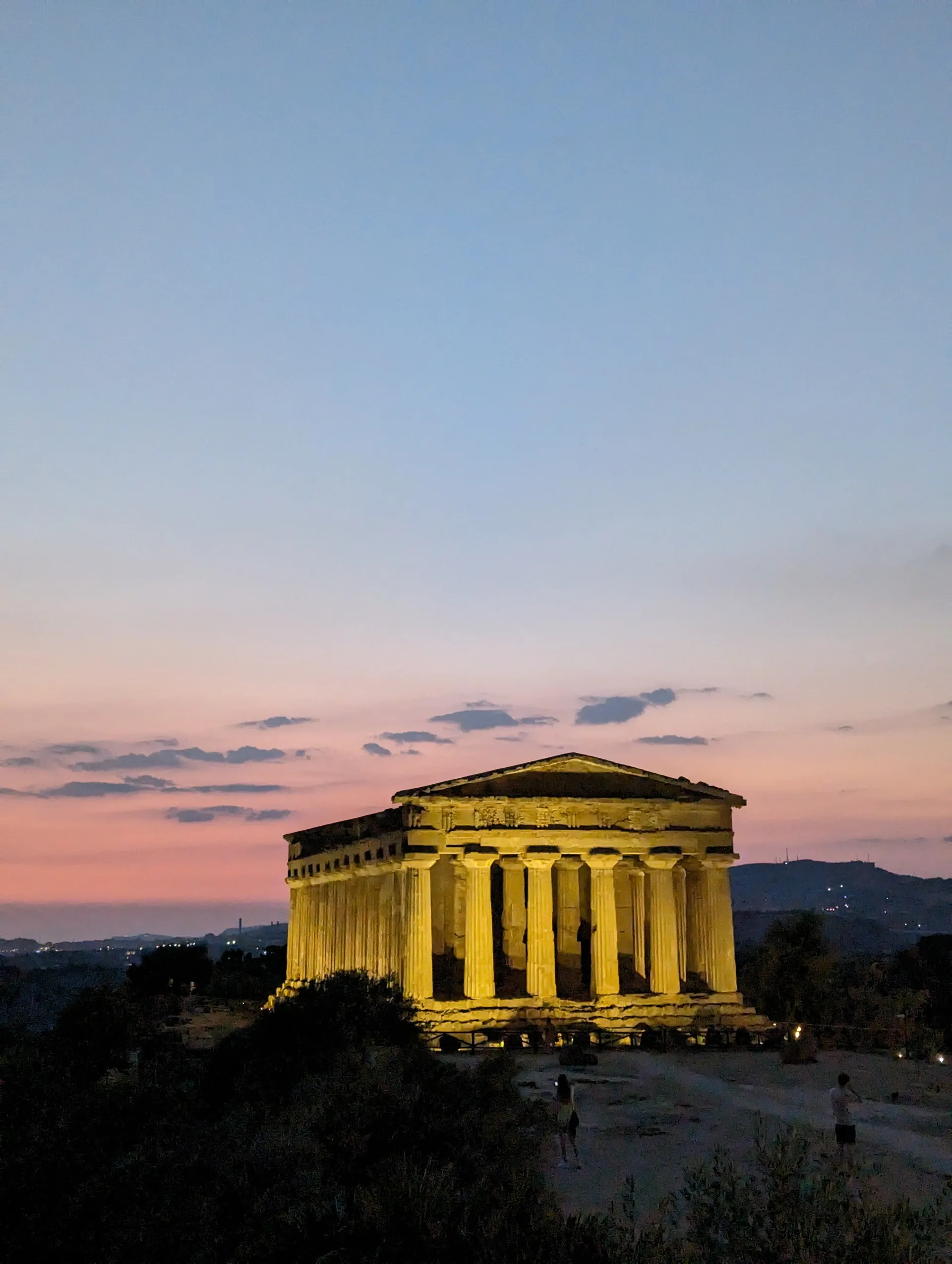 A floodlit Greek temple on a ridge against a pink evening sky.